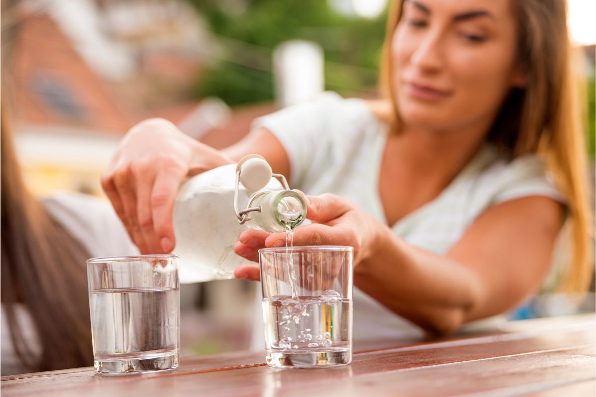 A photo of a woman pouring out water from a glass bottle into glasses.